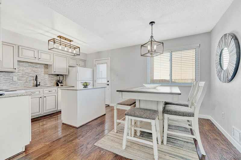 Kitchen with white cabinets, island, and table. Wooden floor. Window with blinds.