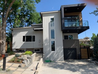 Modern two-story house with light gray siding, black accents, and a glass balcony. Concrete walkway and garden.