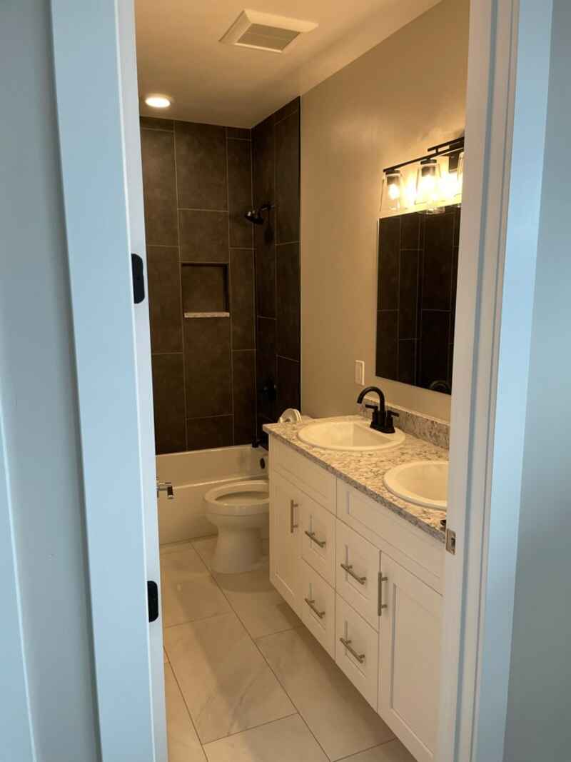 Bathroom with white vanity, dark shower tile, and light-colored flooring. Viewed from doorway.