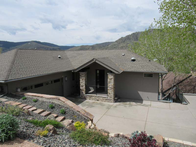 House with brown roof, stone columns, and gray siding, driveway, and mountain backdrop.
