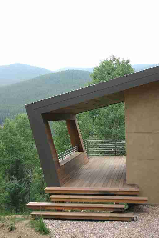 Modern house with a wooden deck and mountain view. Angled roof overhangs the deck; trees in the background.
