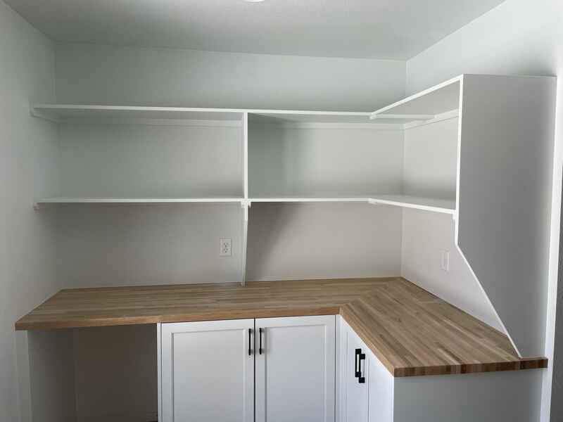 White built-in pantry with wooden countertop, white shelves, and cabinets. Corner setup, empty.