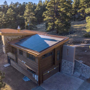 Modern two-story house with solar panels on the roof, set in a wooded area. Wooden exterior, flat roof.