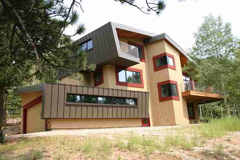 Modern two-story house with slanted roof, brown siding, and tan stucco exterior, surrounded by trees and grass.