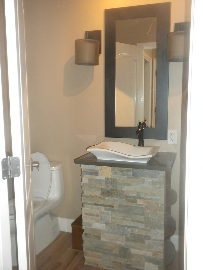 Small bathroom with a stone-faced vanity, vessel sink, mirror, and toilet. Beige walls, brown sconces.