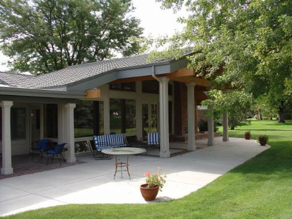 Patio with a concrete surface, featuring pillars and a roof. Blue and white chairs, a table, and potted plants. Green grass and trees.