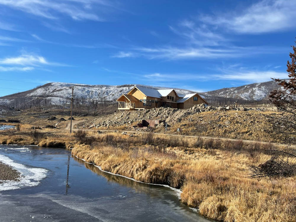 A house on a hill overlooking a stream, mountains in the background, partly cloudy sky.