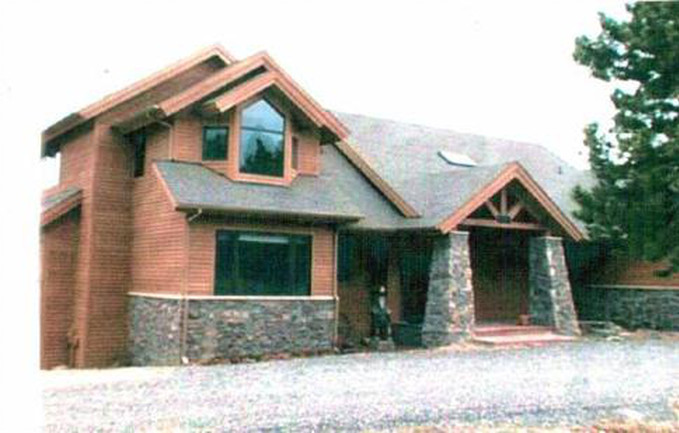 Two-story wooden house with stone accents and a gabled roof, driveway in foreground, trees.