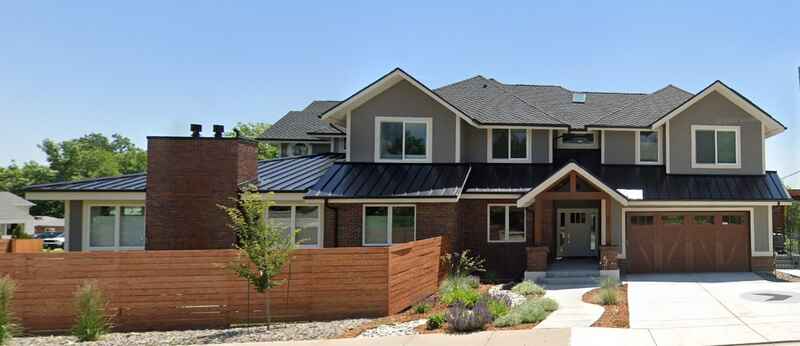 Modern two-story house with gray siding, brown accents, a wooden fence, and a brick chimney on a sunny day.