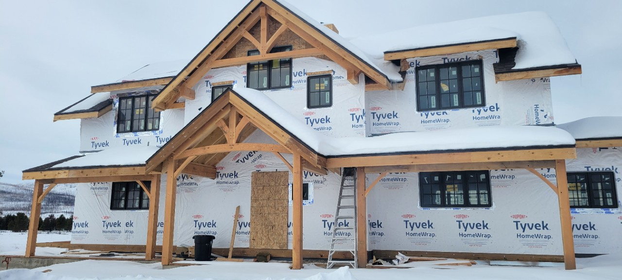 A two-story house under construction, with snow on the roof, white siding, and wooden beams.