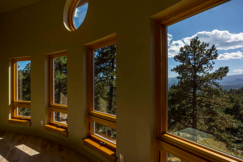 Interior view of a curved room with wooden-framed windows overlooking a forest and mountains under a blue sky.