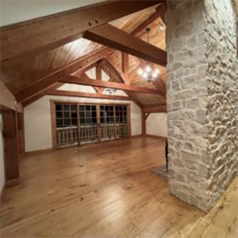 Spacious room with wooden floors, ceiling beams, and a stone fireplace. Large windows in the back.