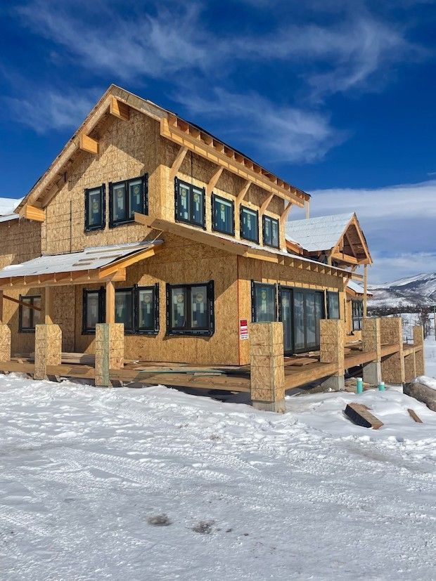 A two-story house under construction with wood framing, black windows, and snow in the foreground against a blue sky.