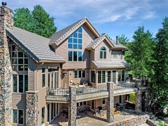 Large beige house with stone accents, large windows, and a wraparound balcony, surrounded by trees under a blue sky.