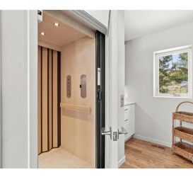 Residential elevator doorway with a cream-colored interior, next to a bathroom with a window and wood floor.