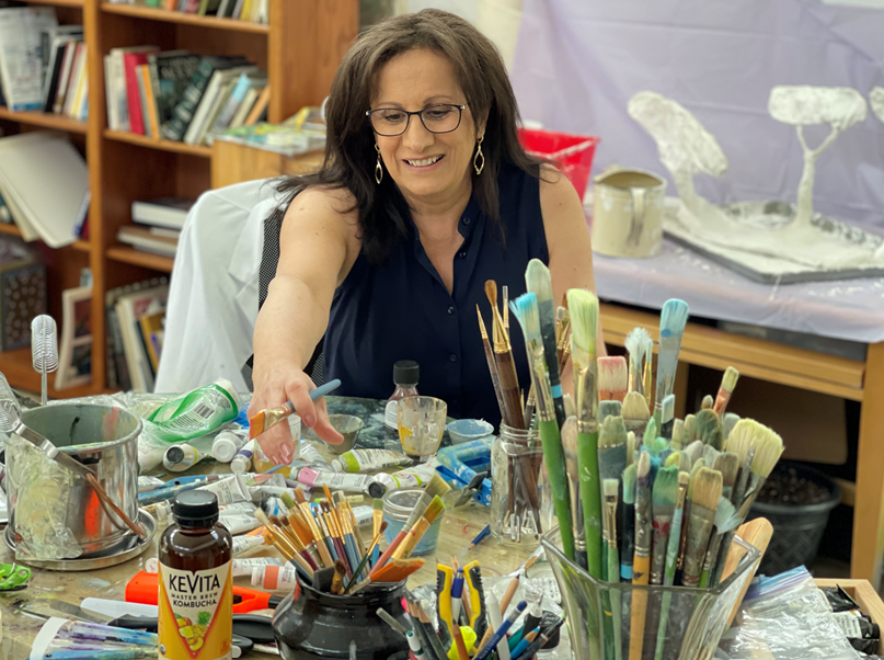 Woman at a cluttered art table reaching for paint. Brushes, paint, and art supplies surround her.