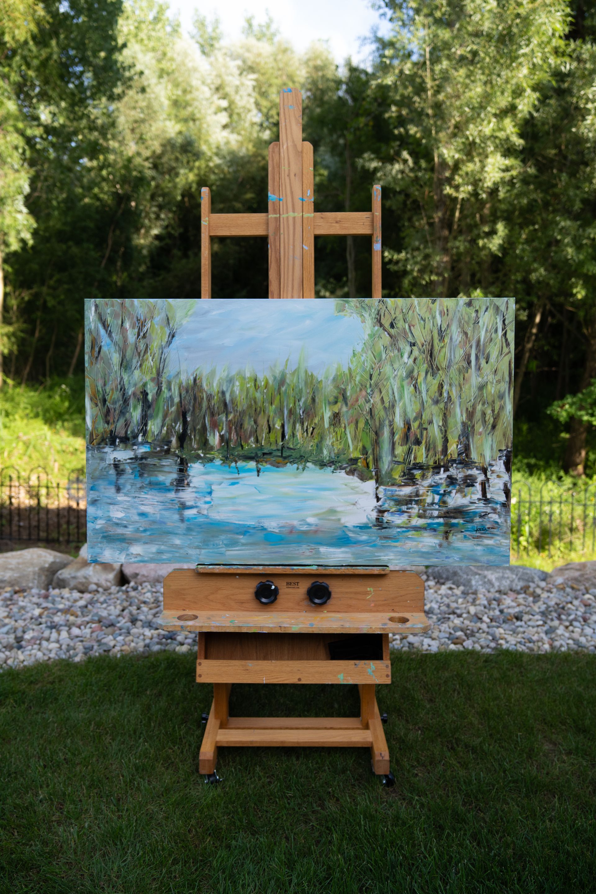Painting on an easel outdoors depicts a blue lake surrounded by green trees.