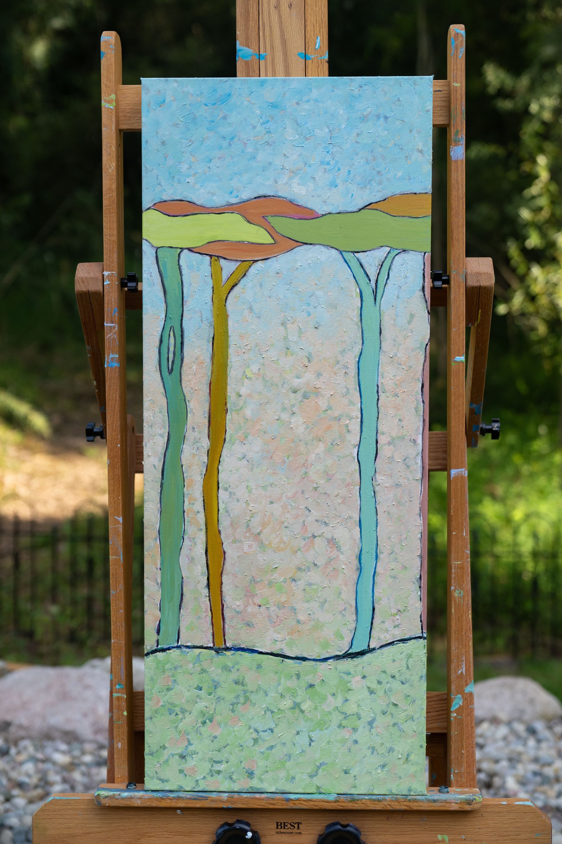 Abstract landscape painting on easel: light blue sky, brown hills, tall green and yellow trees.