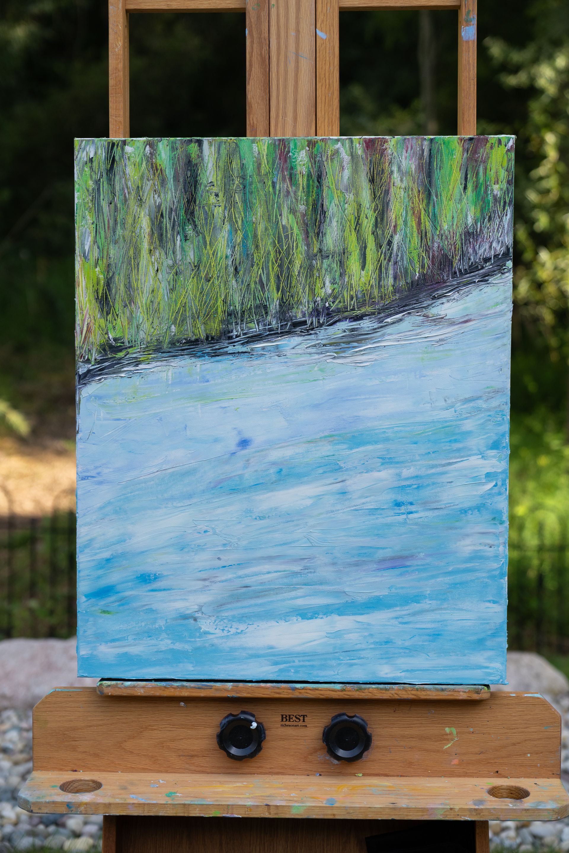 Painting of a blue body of water with a green, textured shoreline on an easel.