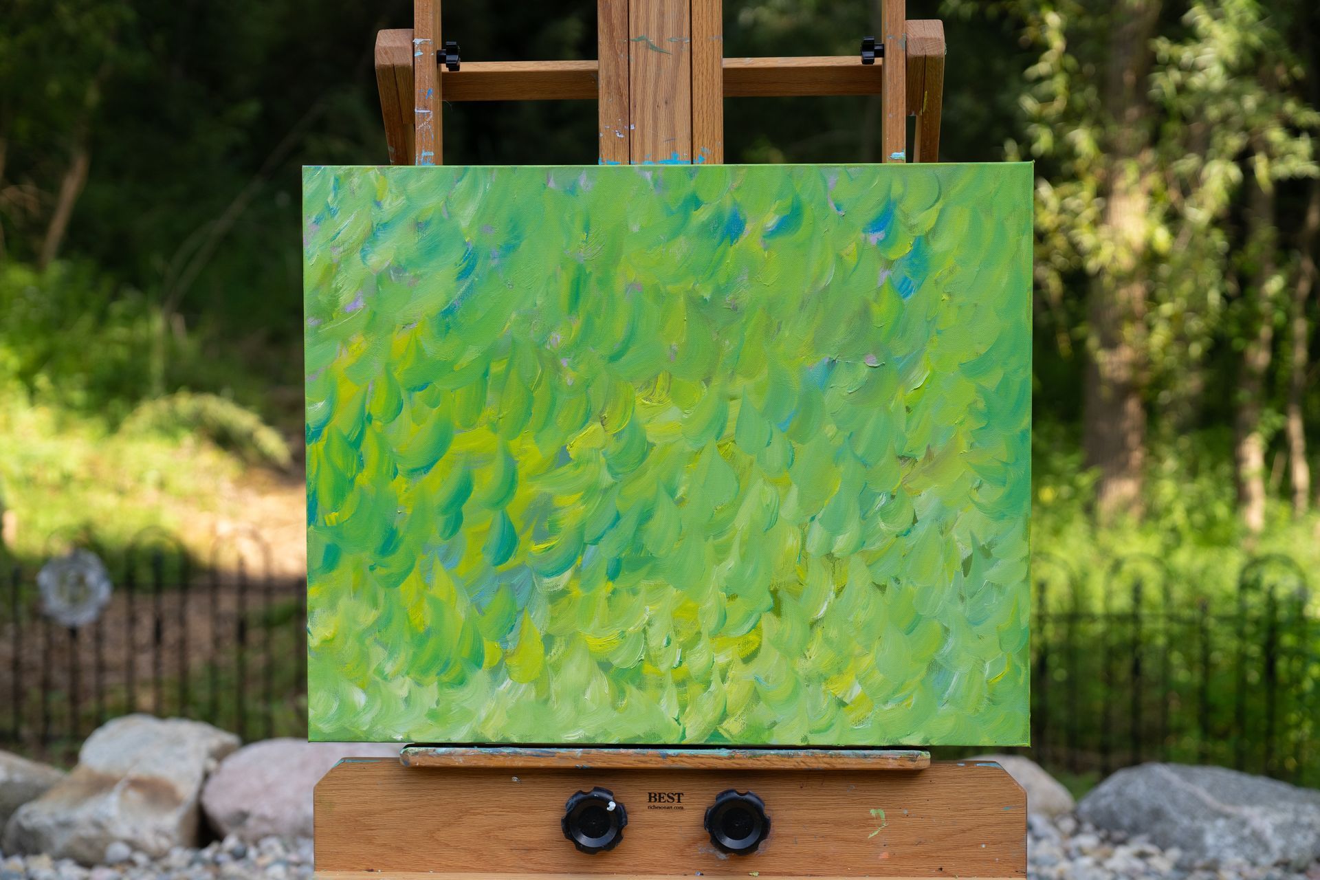 Green and blue abstract painting on a wooden easel outdoors with trees in the background.