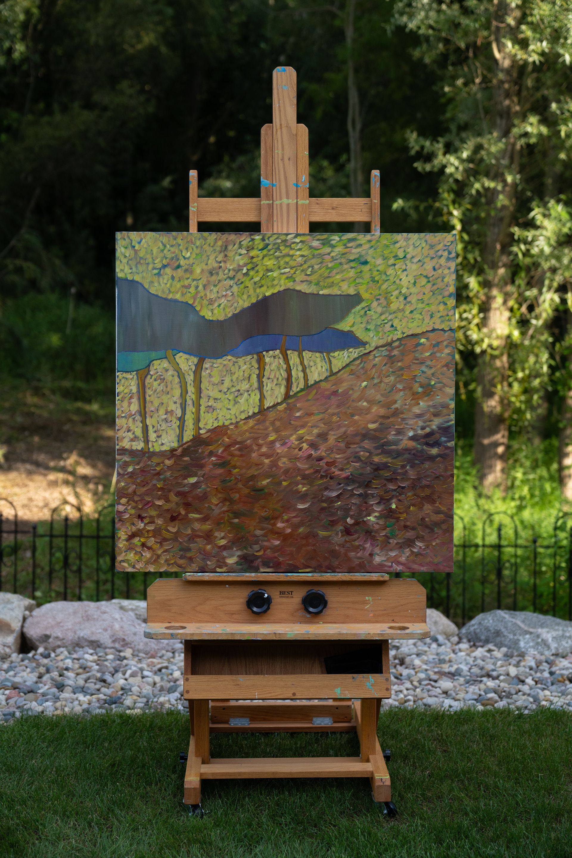 Painting on an easel outdoors. Landscape art with brown, green, blue, and yellow hues. Sunny setting with a wooden easel.