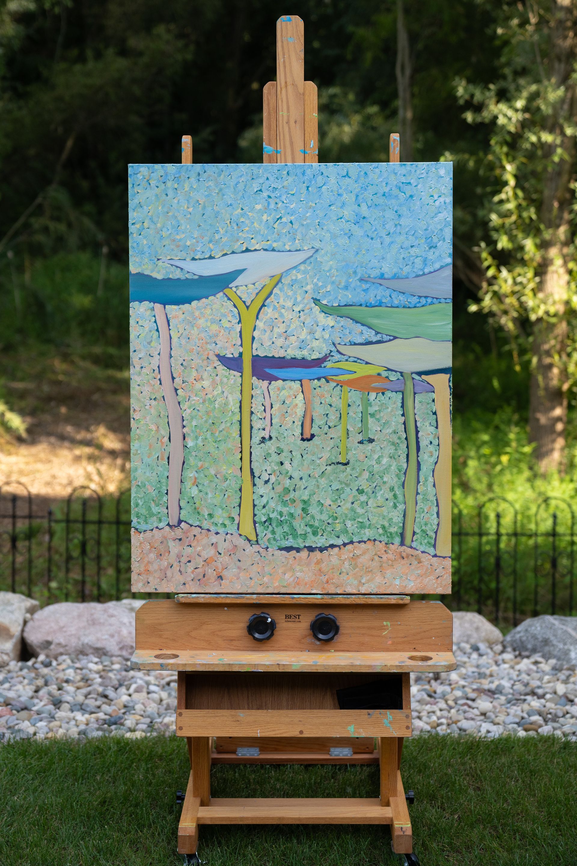 Painting on an easel outdoors: abstract landscape in blue, green, and yellow hues.