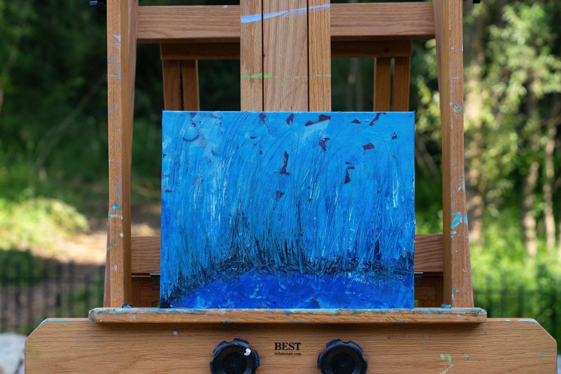 Blue abstract painting on an easel outdoors with blurred green foliage in the background.