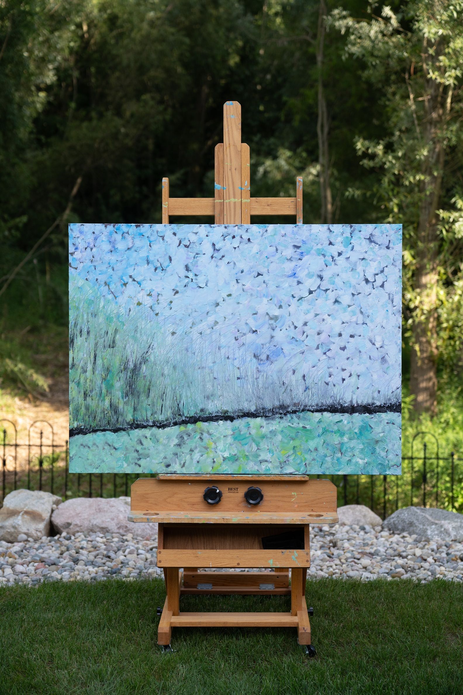 Painting on wooden easel outdoors, depicting a field with a blue sky and sparse, dark details.