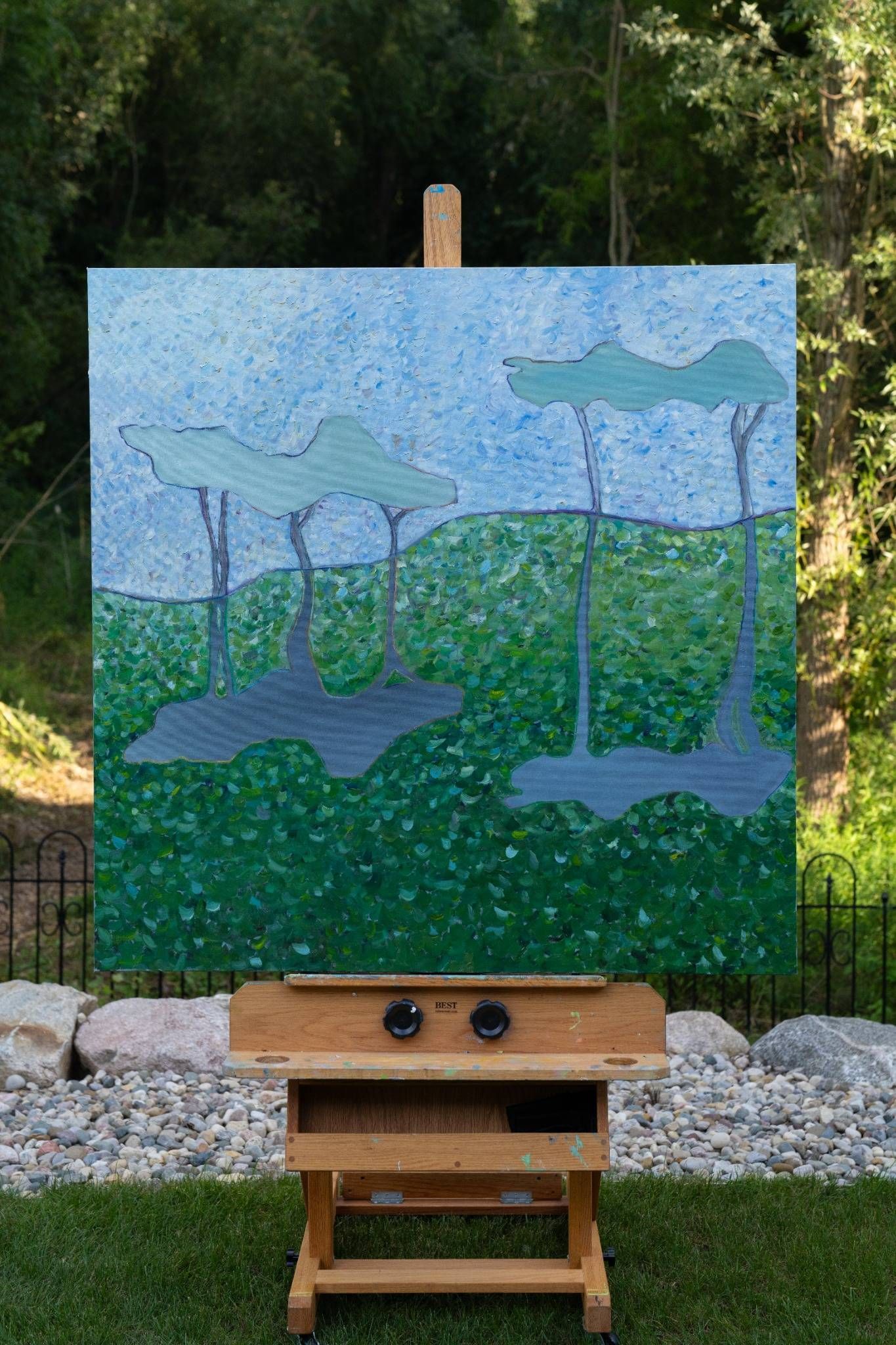 Painting of trees with shadows in a grassy field, displayed on an easel outdoors.