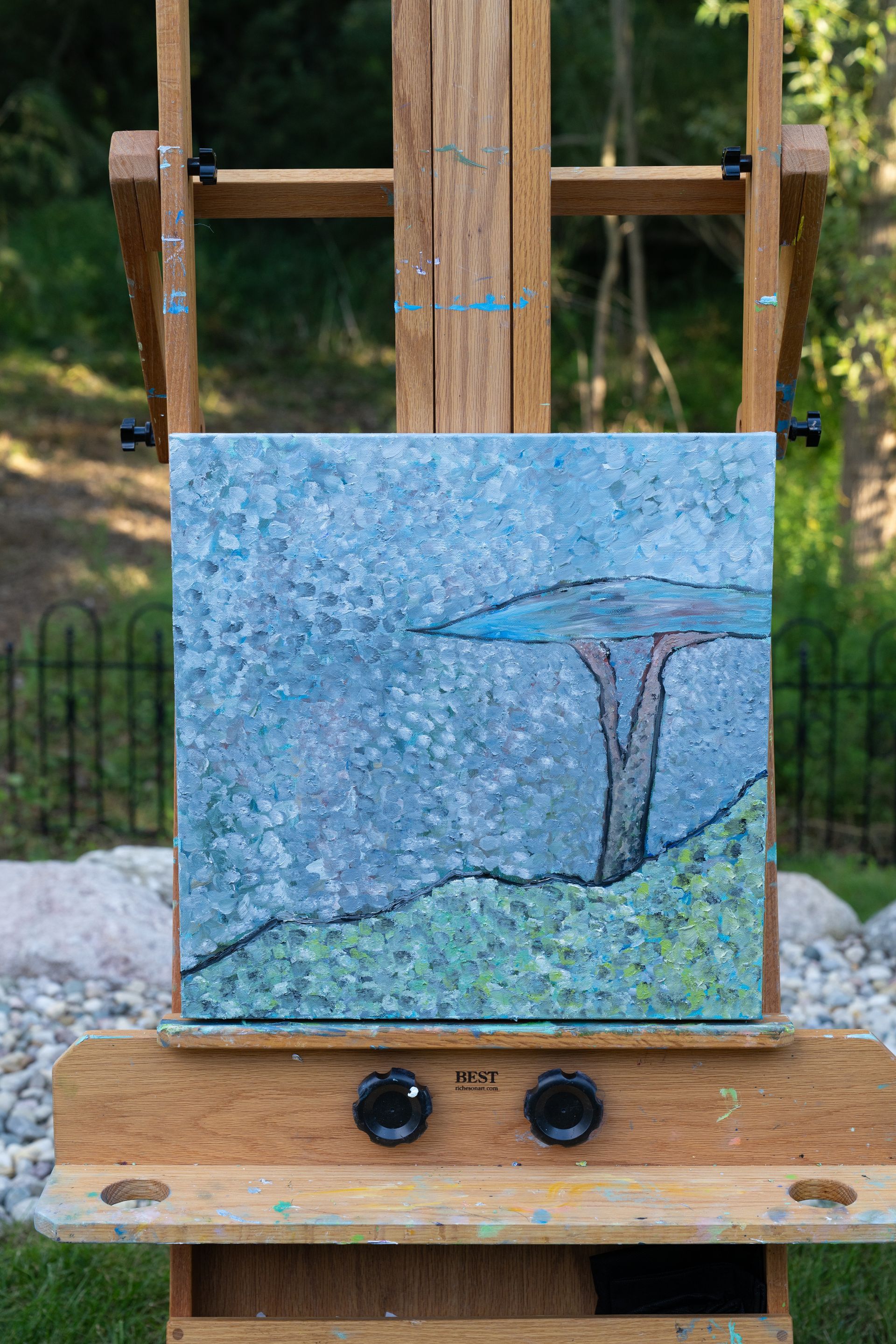 Painting of a blue sky, tree, and green hill, set on a wooden easel outdoors.