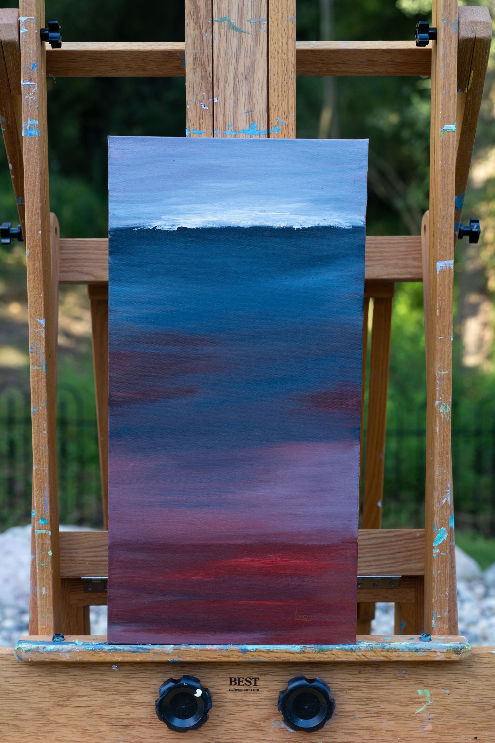 Abstract landscape painting on a wooden easel. Gradient of colors from blue-gray sky to deep red at the bottom.