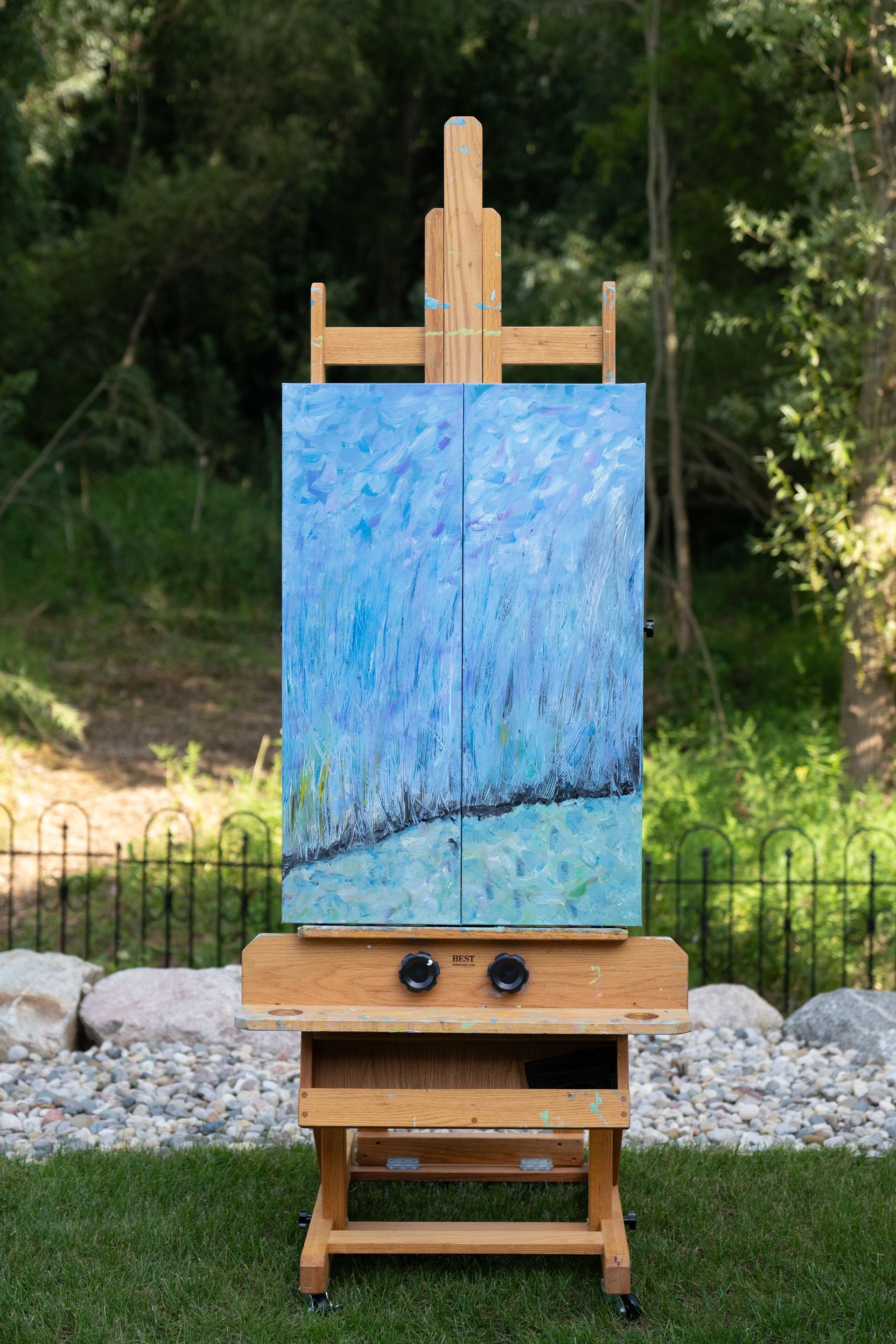 Artwork on easel outdoors, blue abstract painting, wooden easel, green grass background.