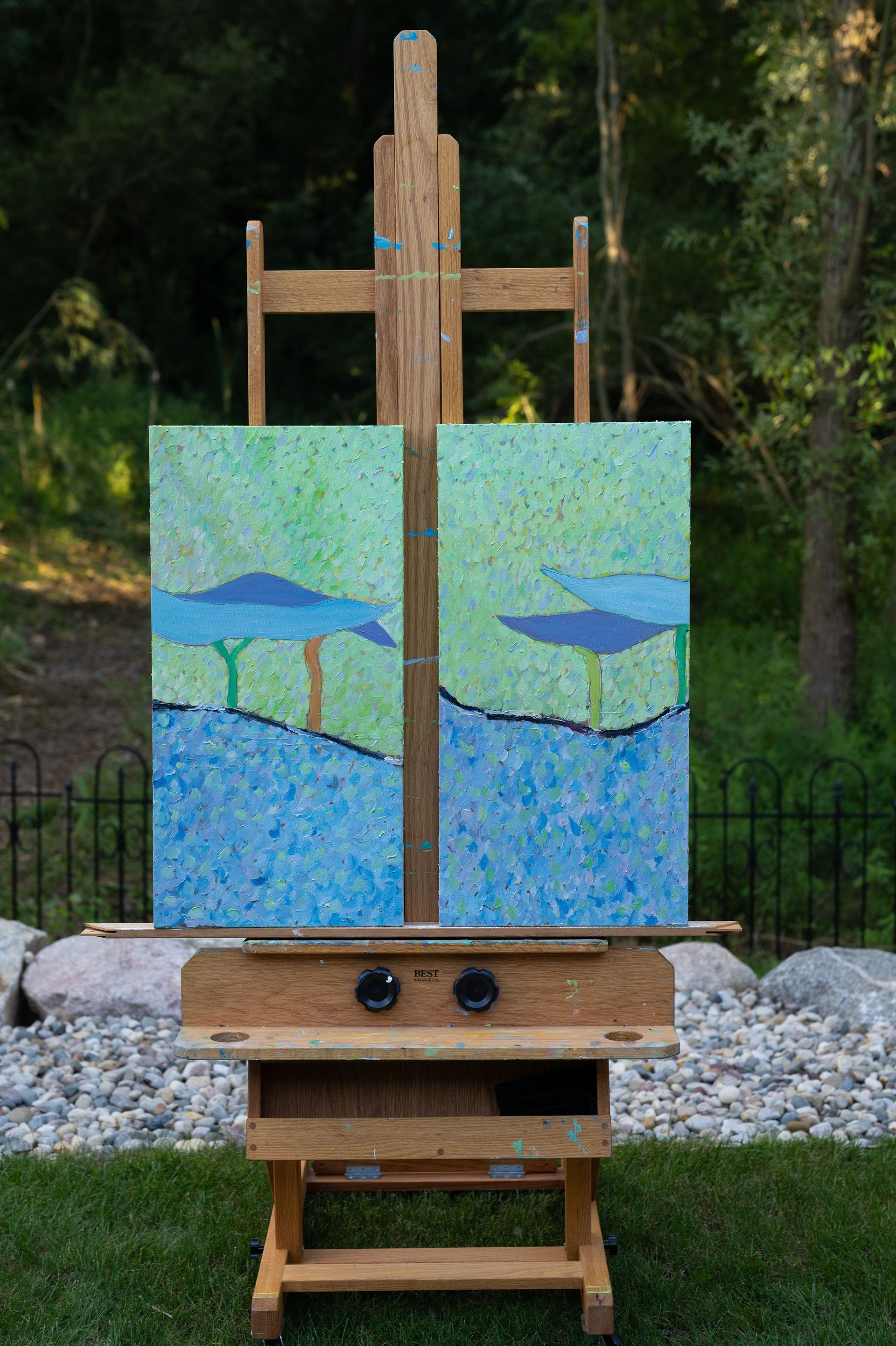 Two abstract paintings on an easel. Blue, green, and brown tones. Trees and water depicted. Outdoors setting.