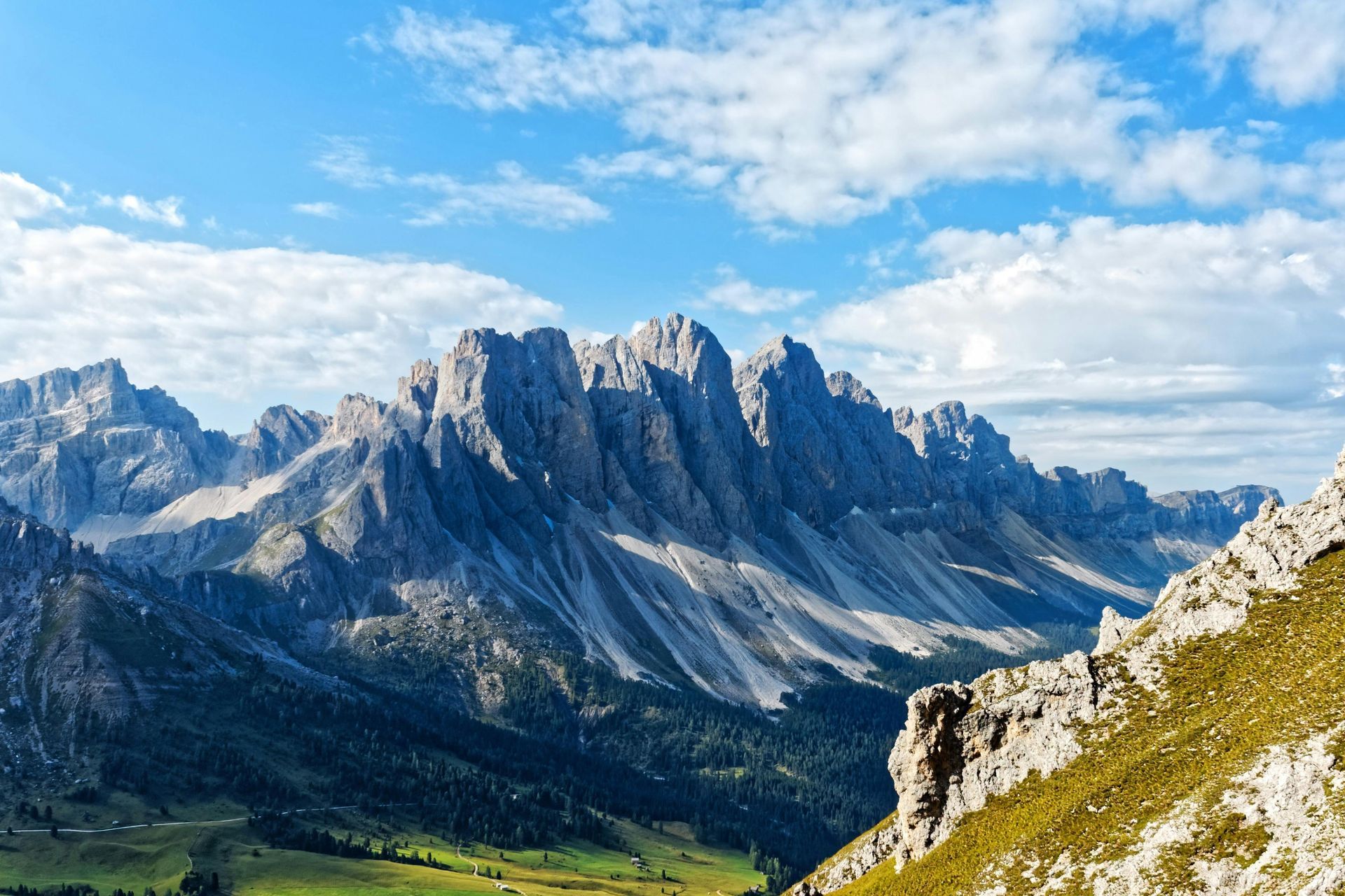 Jagged, gray mountain range under a partly cloudy, blue sky. Green valley in foreground.