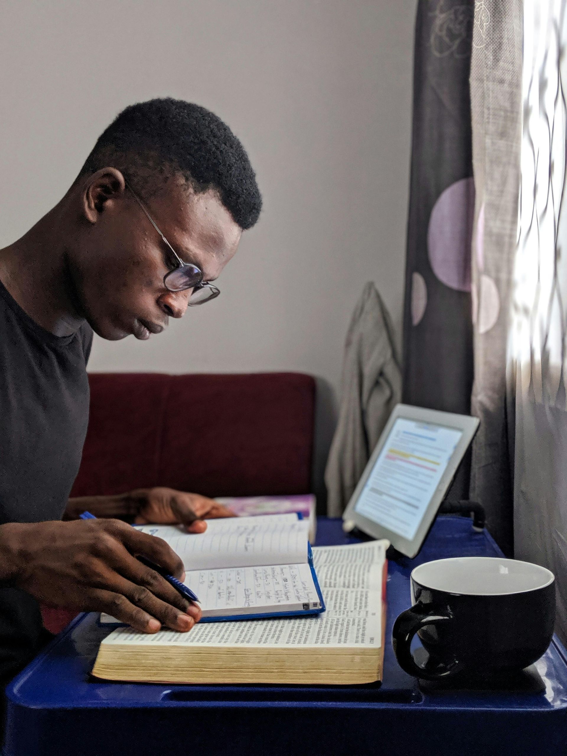Person studying a Bible on a table with a tablet and journal and mug
