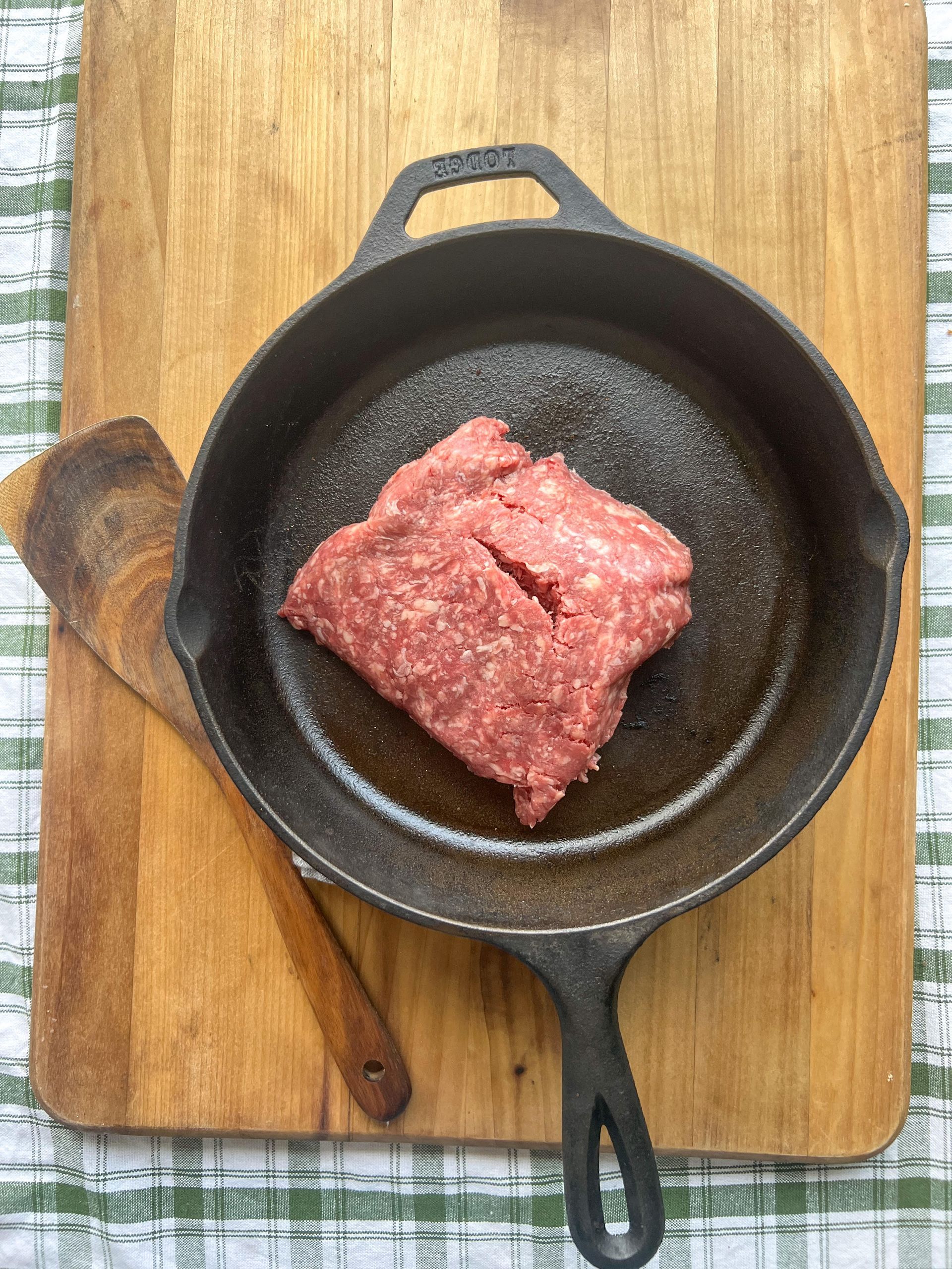 A piece of ground beef is in a cast iron skillet on a wooden cutting board.