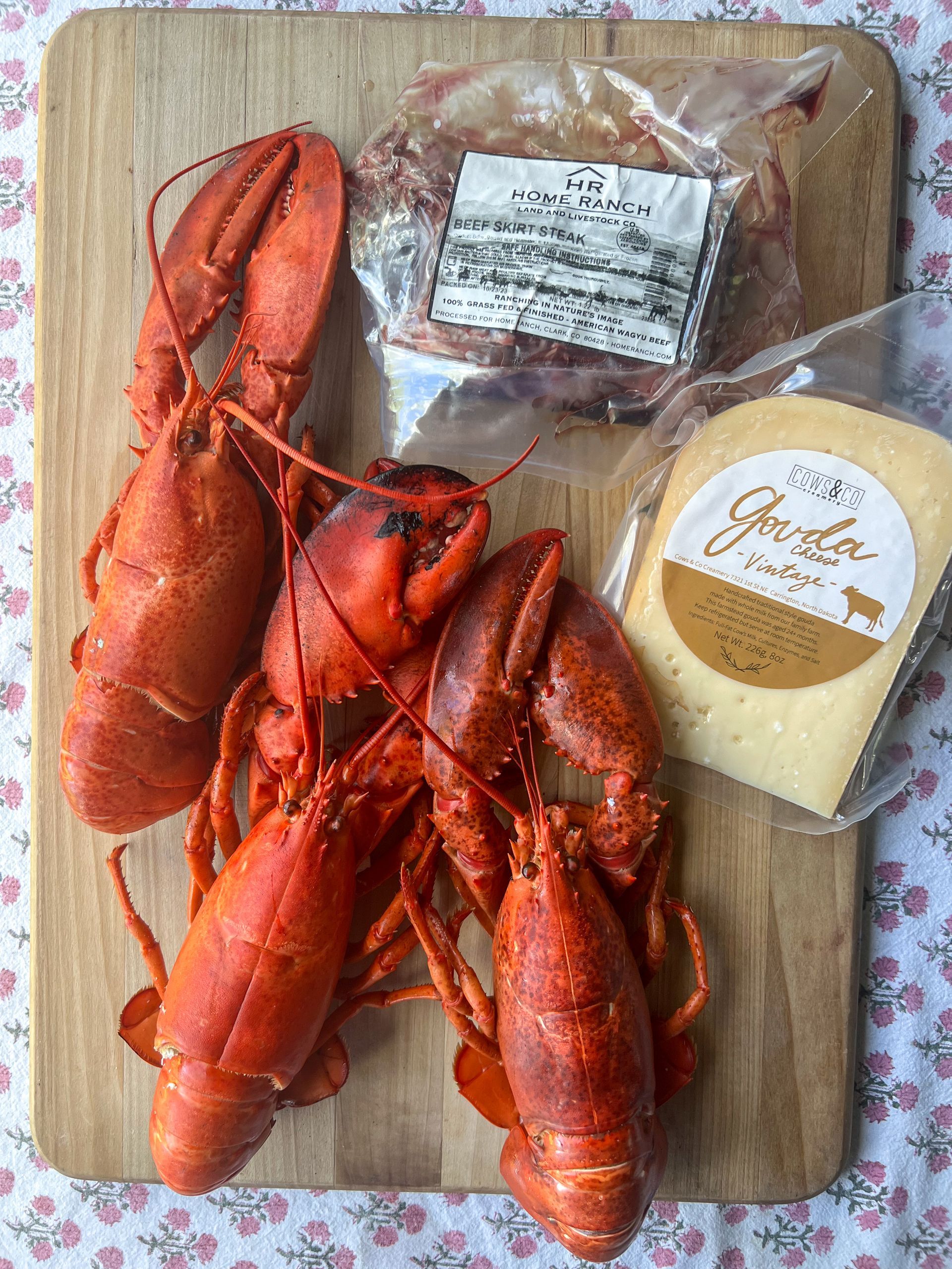 Two lobsters and a piece of cheese are on a wooden cutting board.