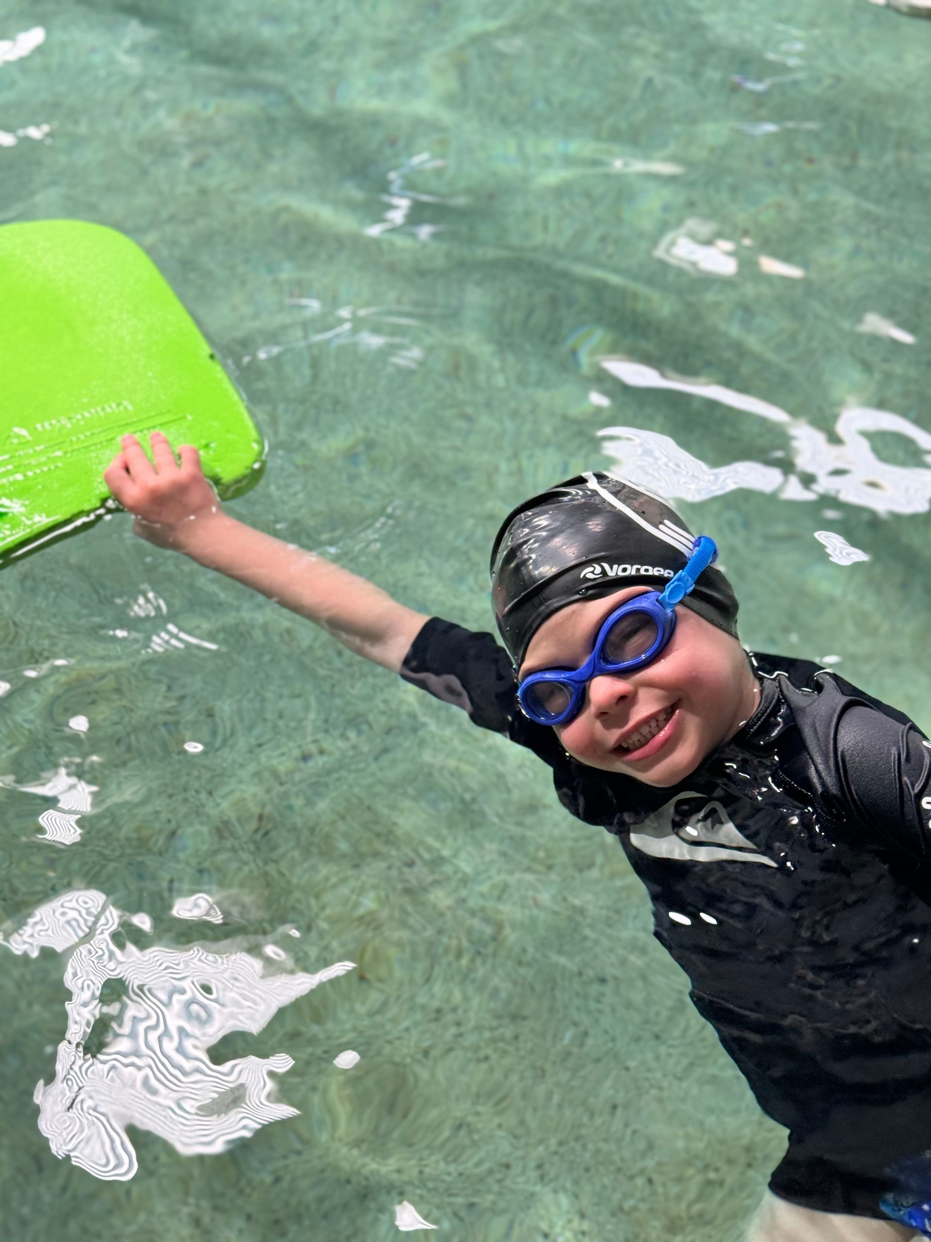 A young boy wearing goggles and a swim cap is holding a green boogie board in the water.