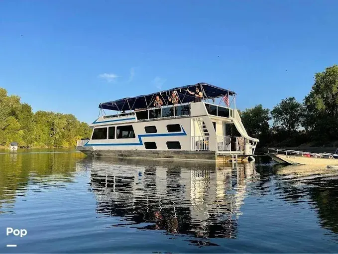 Houseboat on water with people on top deck, trees in background.