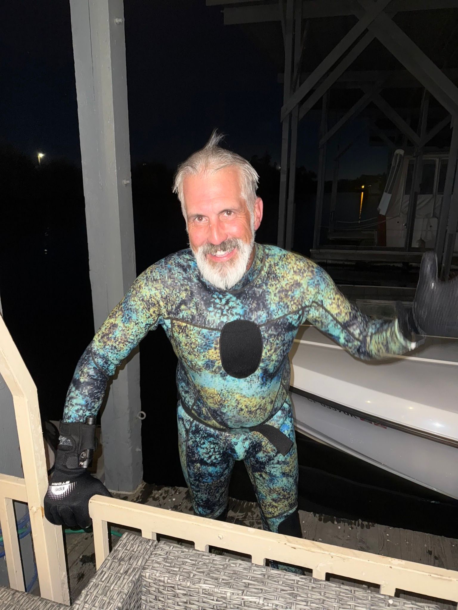 Man in wetsuit stands beside boat, smiling. Gray beard, hair spiked up. Outdoors, evening setting.