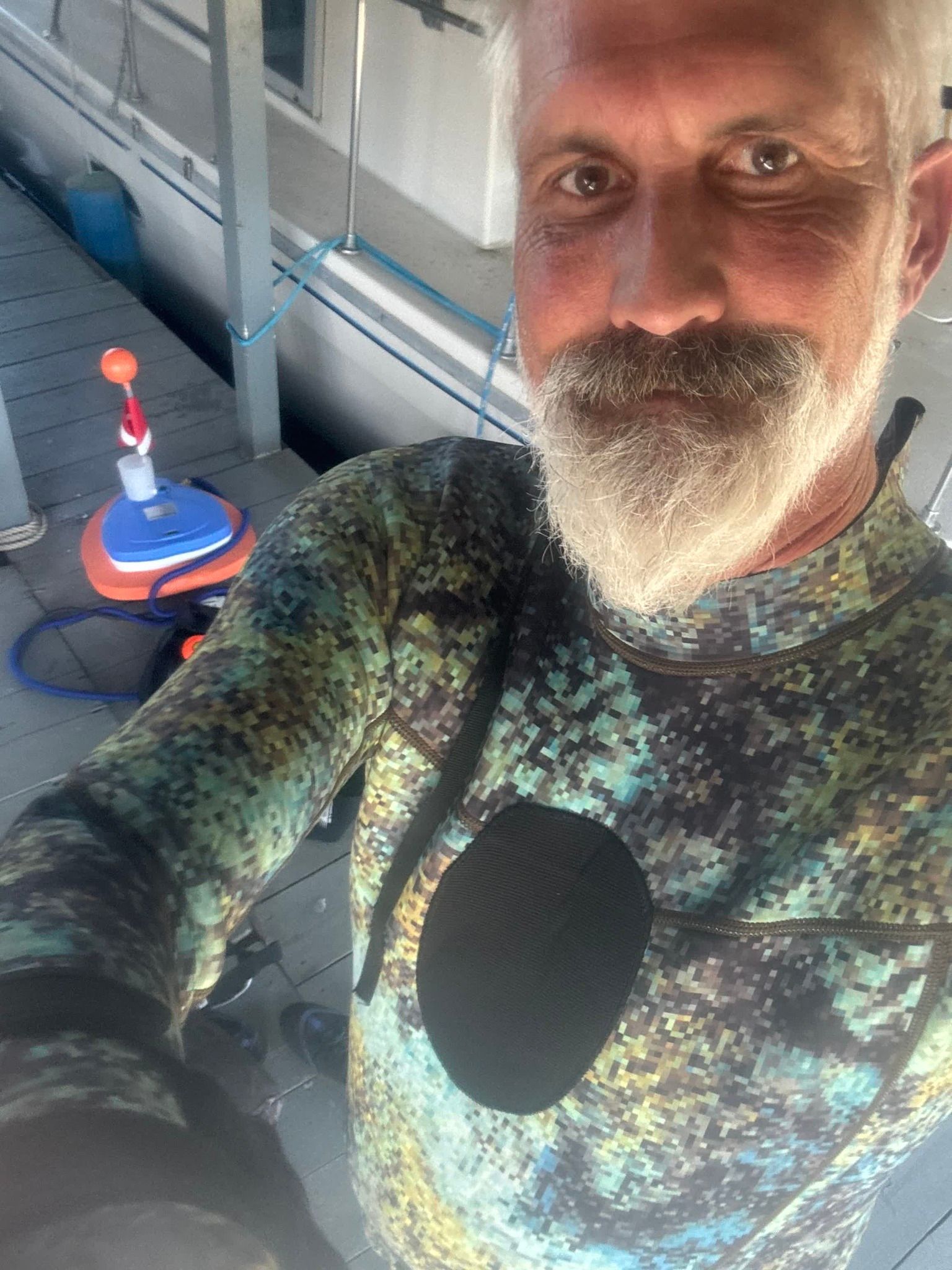 Man with gray beard in wetsuit on dock, looking at camera.