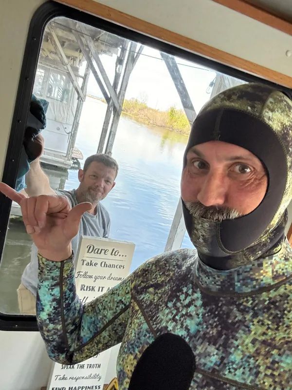 Man in dive suit gives shaka; another man in background. Outdoors, next to water.