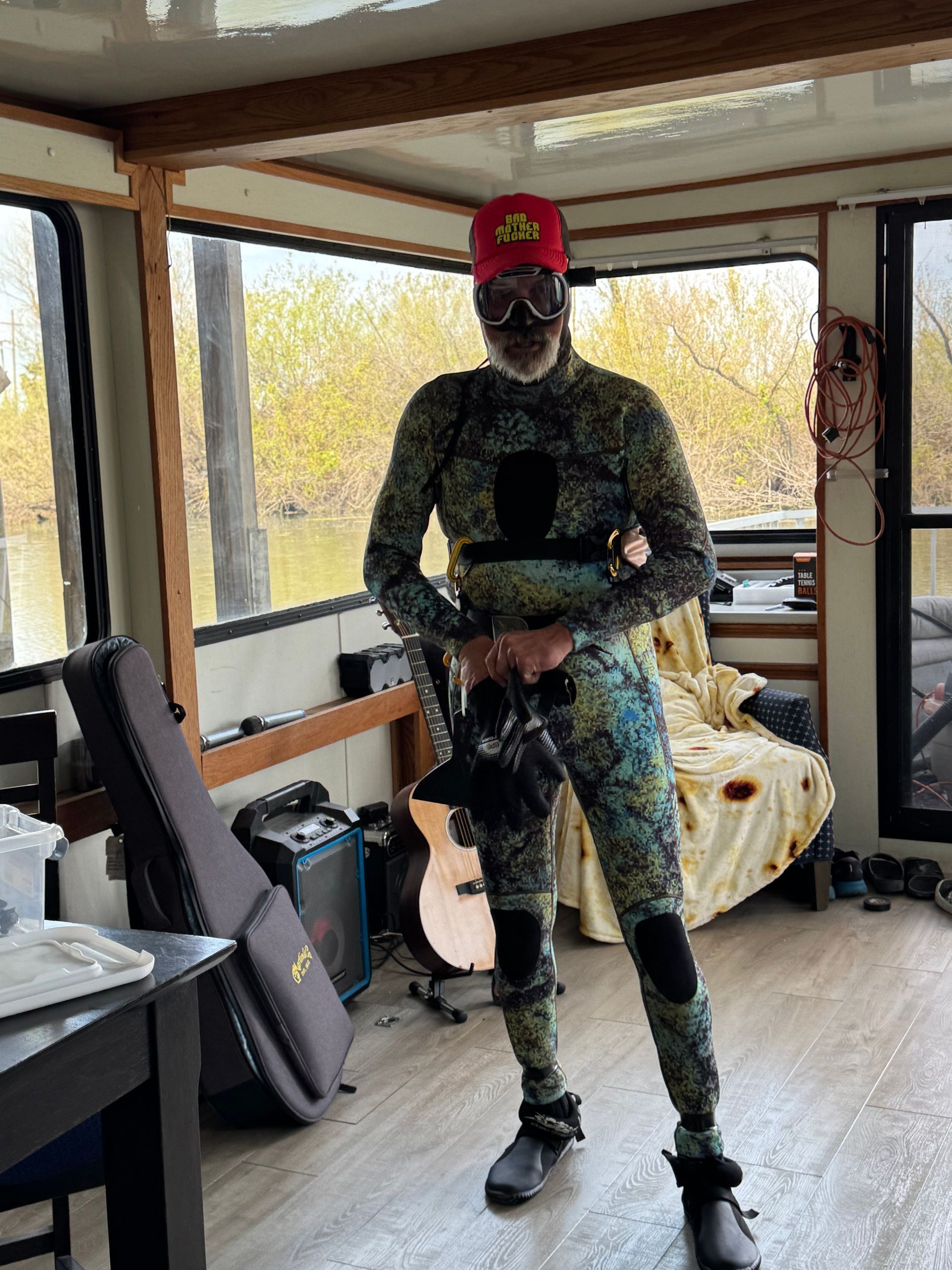 Man in wetsuit, cap, mask holding diving gear, inside boat, river view.
