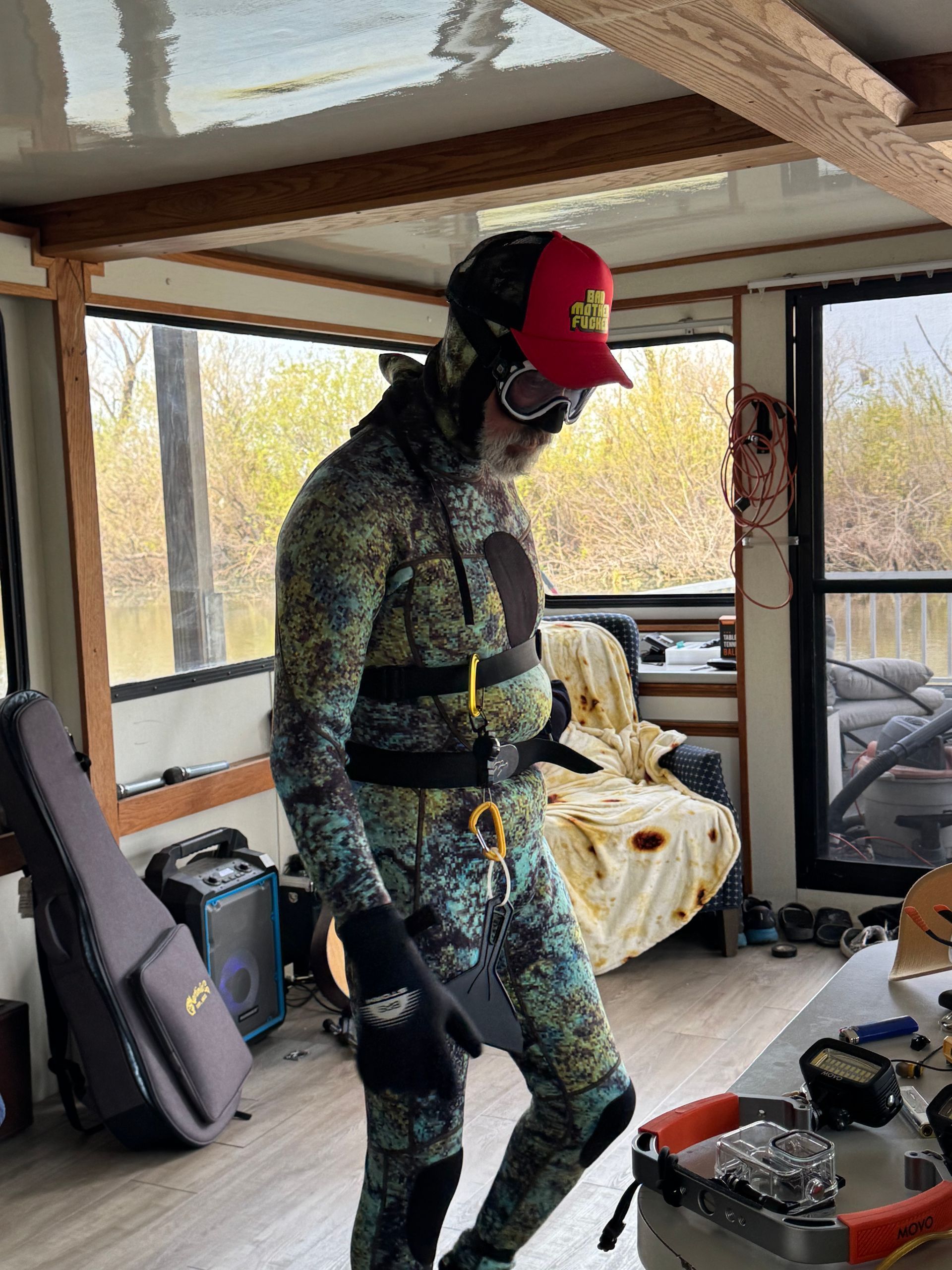Person in camouflage wetsuit, hat, and goggles indoors; appears to be preparing for diving.
