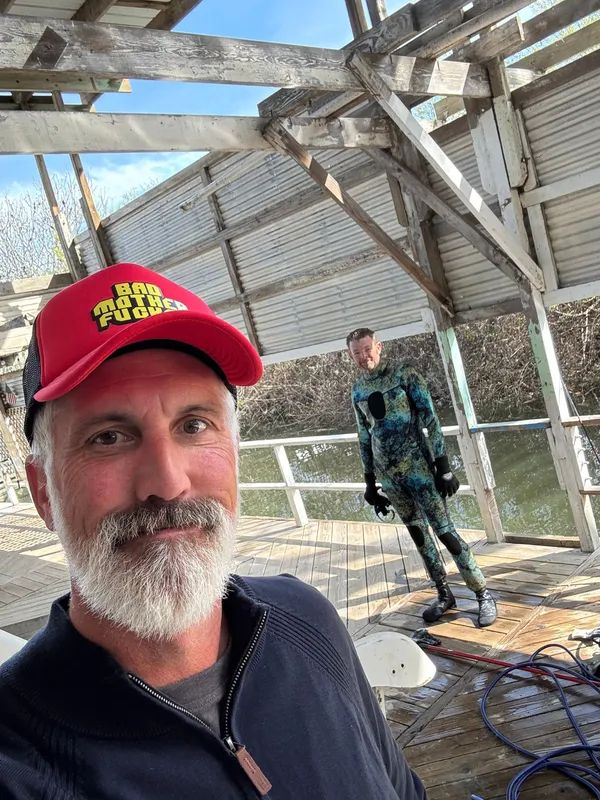 Man in red hat takes selfie, another in camo wetsuit stands nearby on a dock.