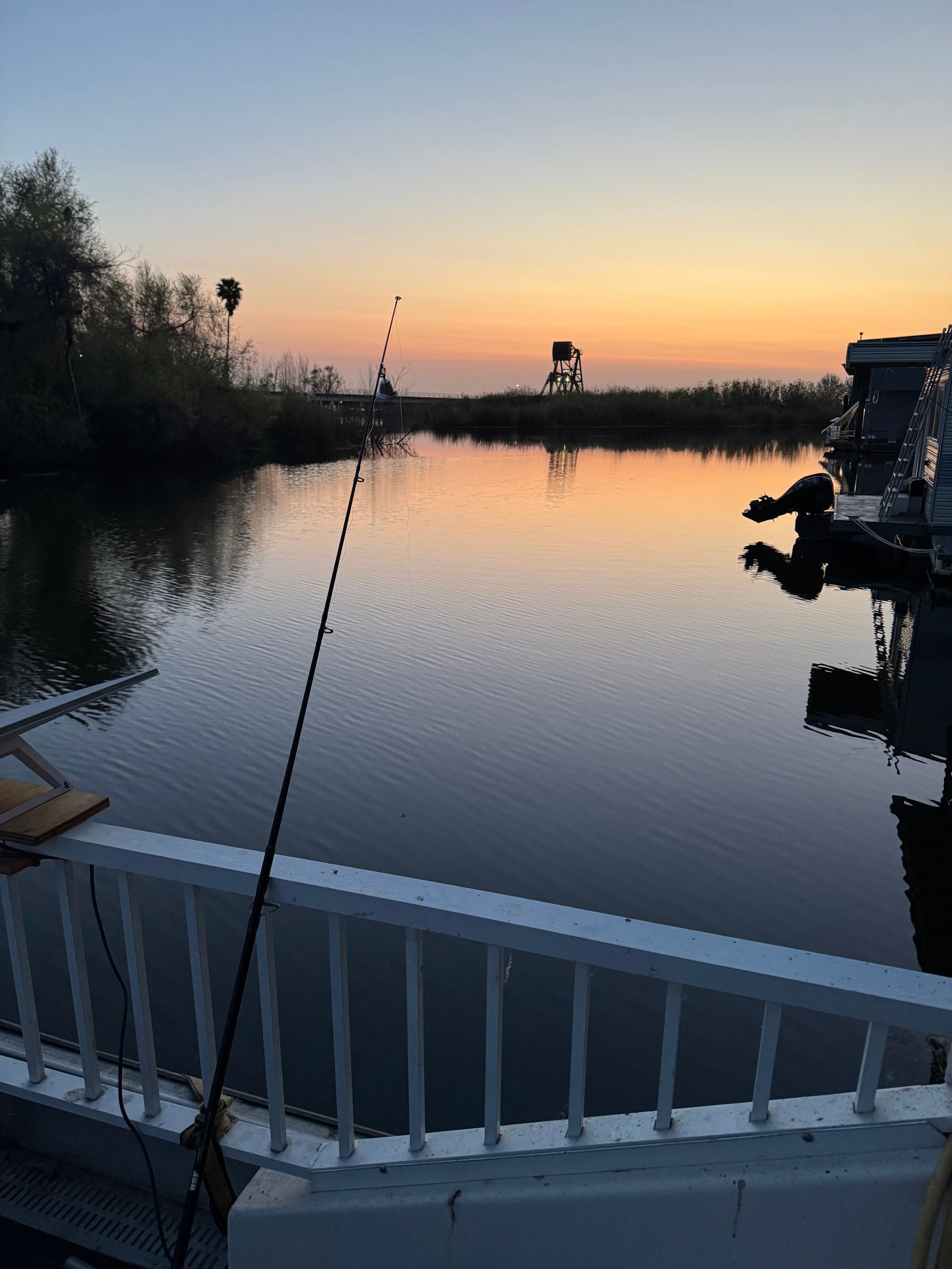 Fishing rod on a deck overlooking calm water at sunset with a muted orange and blue sky.
