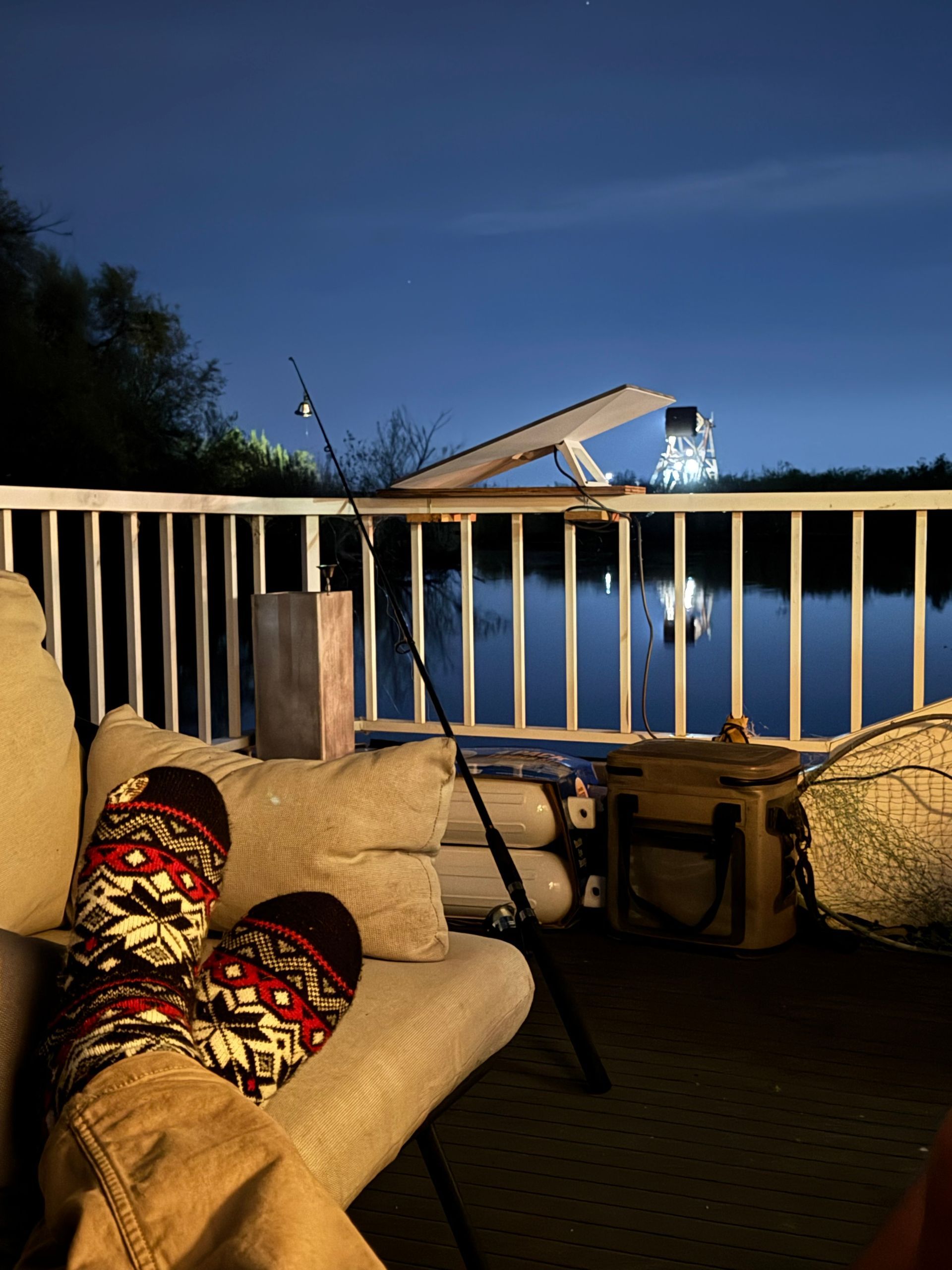 Person wearing patterned socks on a porch at night, fishing rod, water, lit structure in the background.