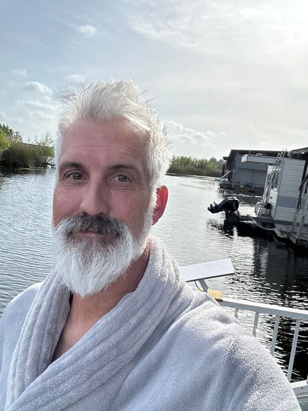 Man with gray beard in robe by water, sunlit, with houseboats in background.
