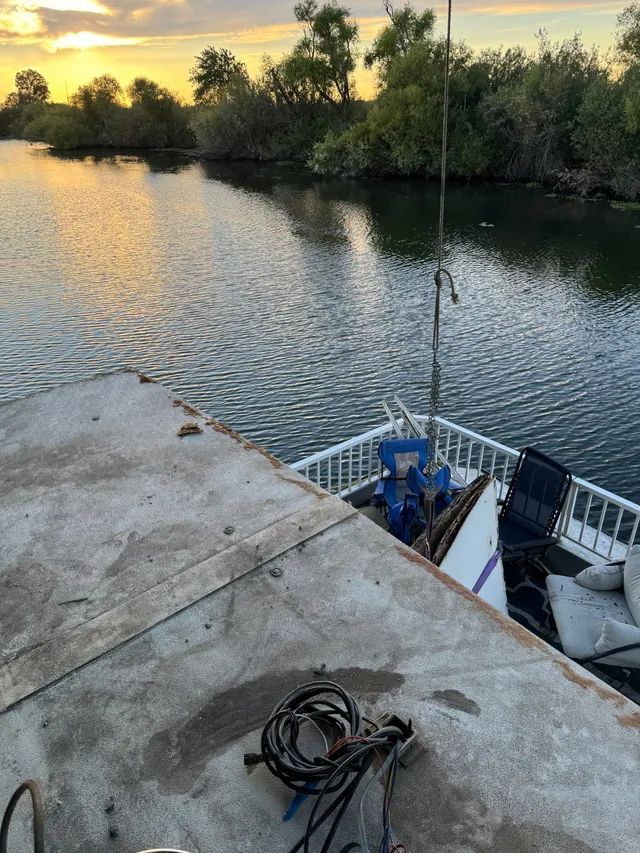 A boat's deck overlooks a waterway at sunset. White railings surround the boat's bow, with a mast and miscellaneous items.