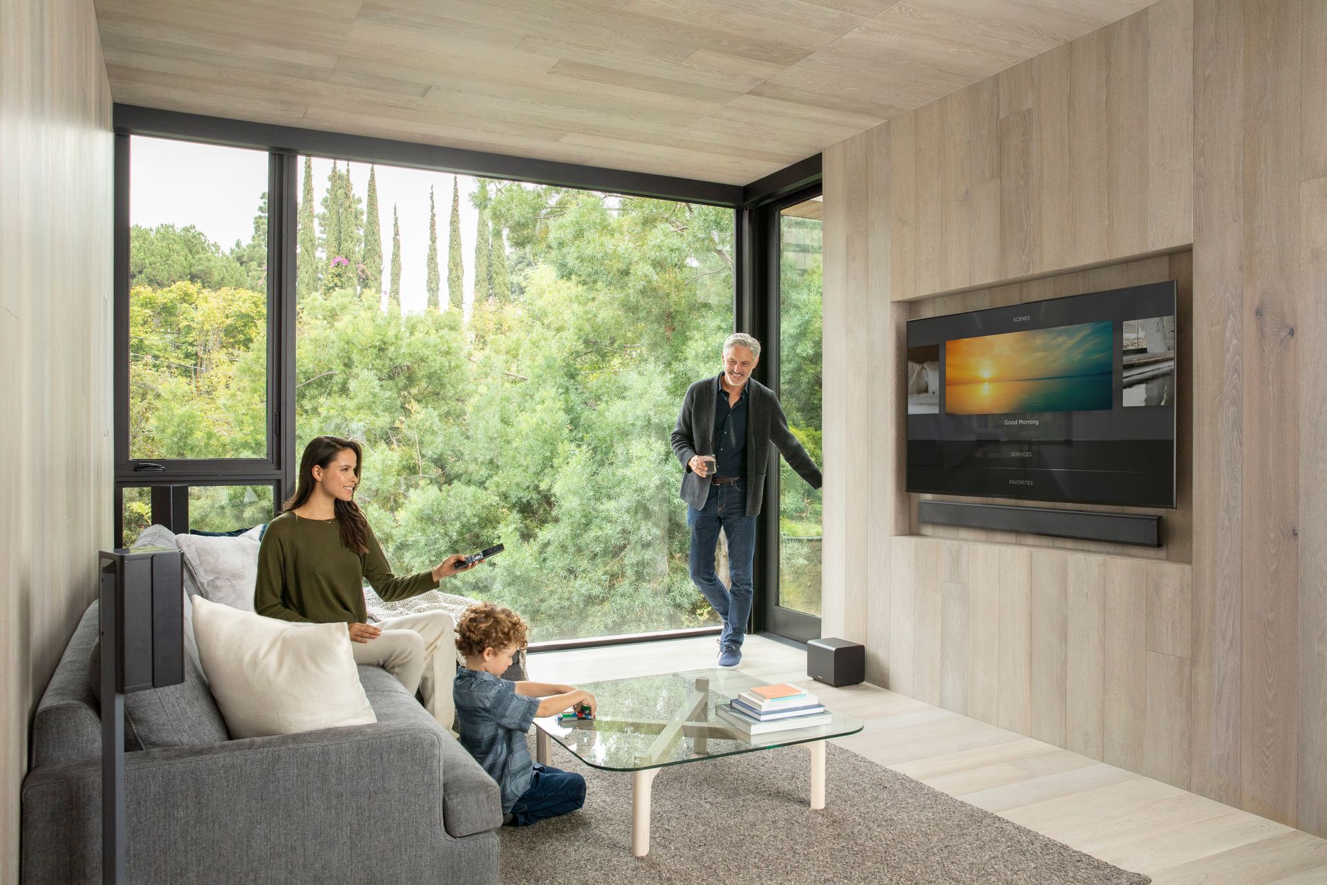 Family watches TV in modern living room with large window and forest view.